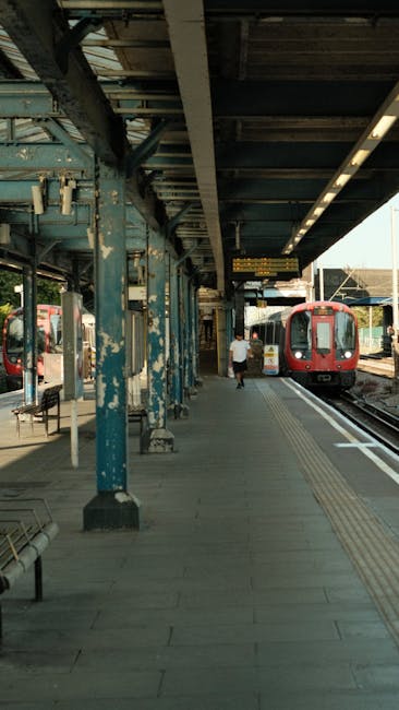 Ilford station platform with a glass and steel roof overhead, exposed brick walls on either side, and tactile yellow safety lines on the wet, textured surface. A train is arriving in the background, w