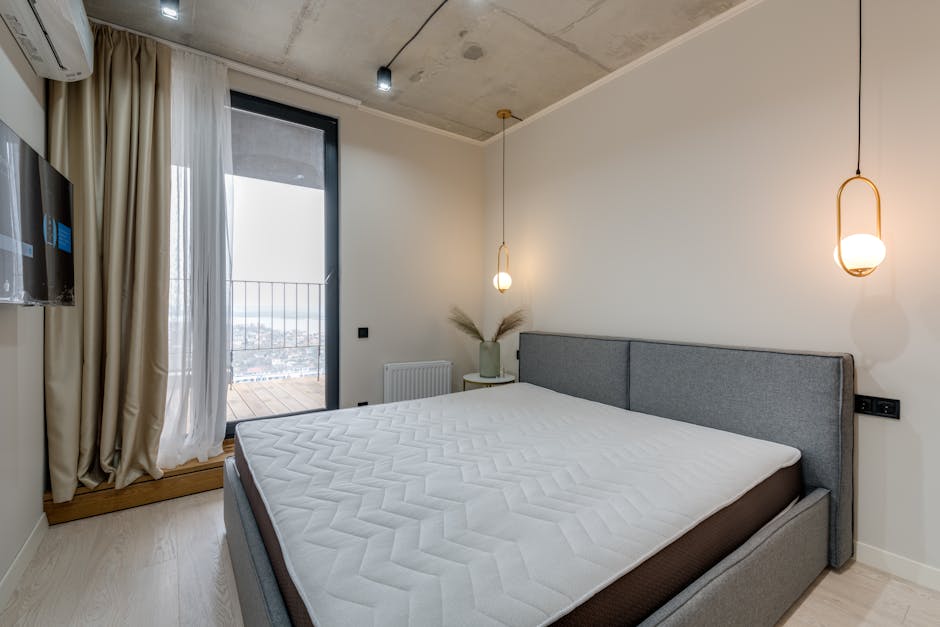 A minimalist bedroom featuring a grey upholstered bed with a blank white mattress, situated against a white wall. To the left, a large glass door with beige curtains leads to a balcony, allowing natur