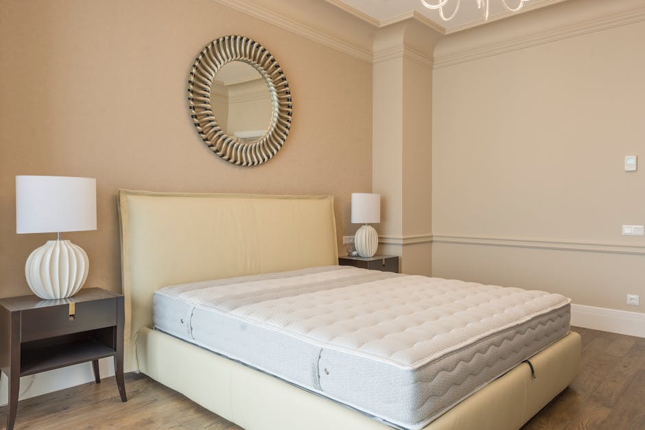 A minimalist bedroom featuring a grey upholstered bed with a blank white mattress, situated against a white wall. To the left, a large glass door with beige curtains leads to a balcony, allowing natur